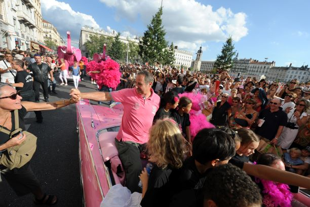Photo de Guy Darmet en T-shirt type polo rose dans une voiture (Cadillac) rose, entouré de jeunes, saluant le public au passage lors du Défilé de la Biennale de la Danse en 2010, place Bellecour. En arrière plan : grand public et éléphant rose.
