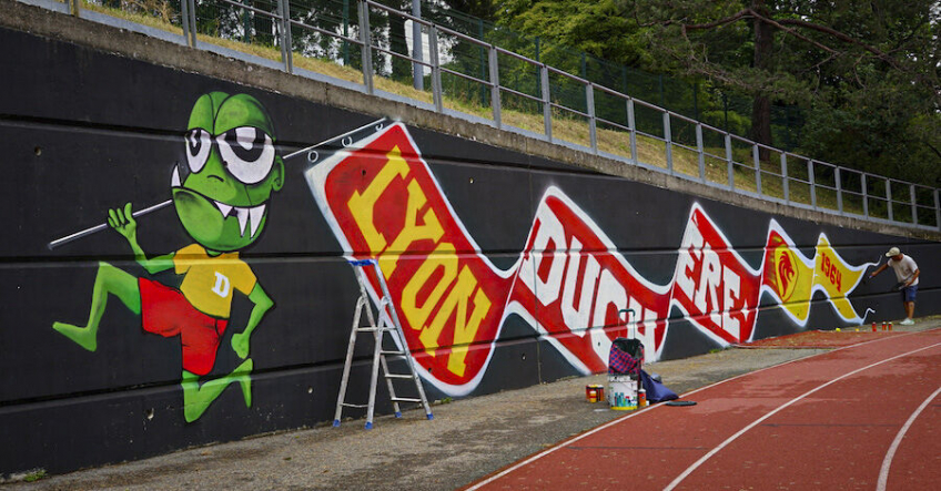 Photo d'un graffiti d'une créature verte contente en train de courir à côté de la piste du stade La Duchère, portant un drapeau "Lyon Duchère 1964"